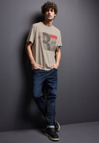 Beige grafisk t-shirt med en stiliserad bokstav "R" och abstrakt design. I kombination med blå jeans och svarta skor med vita sulor.