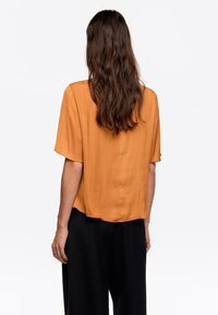 Pomarańczowa bluzka z krótkim rękawem o gładkiej fakturze i zaokrąglonym dole, zestawiona z czarnymi spodniami o szerokich nogawkach. Widok od tyłu.