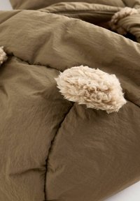 Brun pufferstoff med en rynkig textur, med mjuka beige pom-poms som detalj på kanterna.