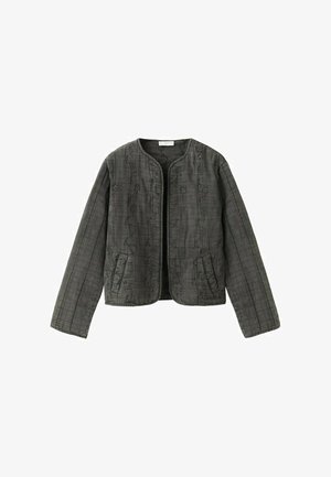 Ung kvinde står iført en gennemsigtig beige top, en mørk tekstureret jakke, grå vidde-benet jeans, brune sko og en rød choker halskæde mod en hvid væg.