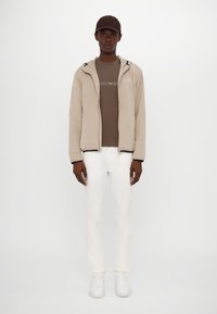 Beige Steppjacke mit schwarzen Reißverschlussdetails, braunes eng anliegendes Shirt, weiße Jeans und weiße Sneaker. Das Model trägt eine braune Mütze.