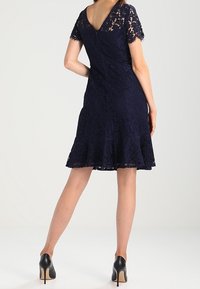 Donna in piedi con le spalle rivolte alla telecamera, che indossa un vestito di pizzo blu navy con maniche corte e scarpe con tacco nero su uno sfondo bianco.