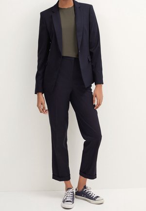 Person trägt einen marineblauen Blazer und passende kurze Hosen mit umgeschlagenen Säumen, ein olivgrünes T-Shirt und marineblaue Sneakers mit weißen Schnürsenkeln.