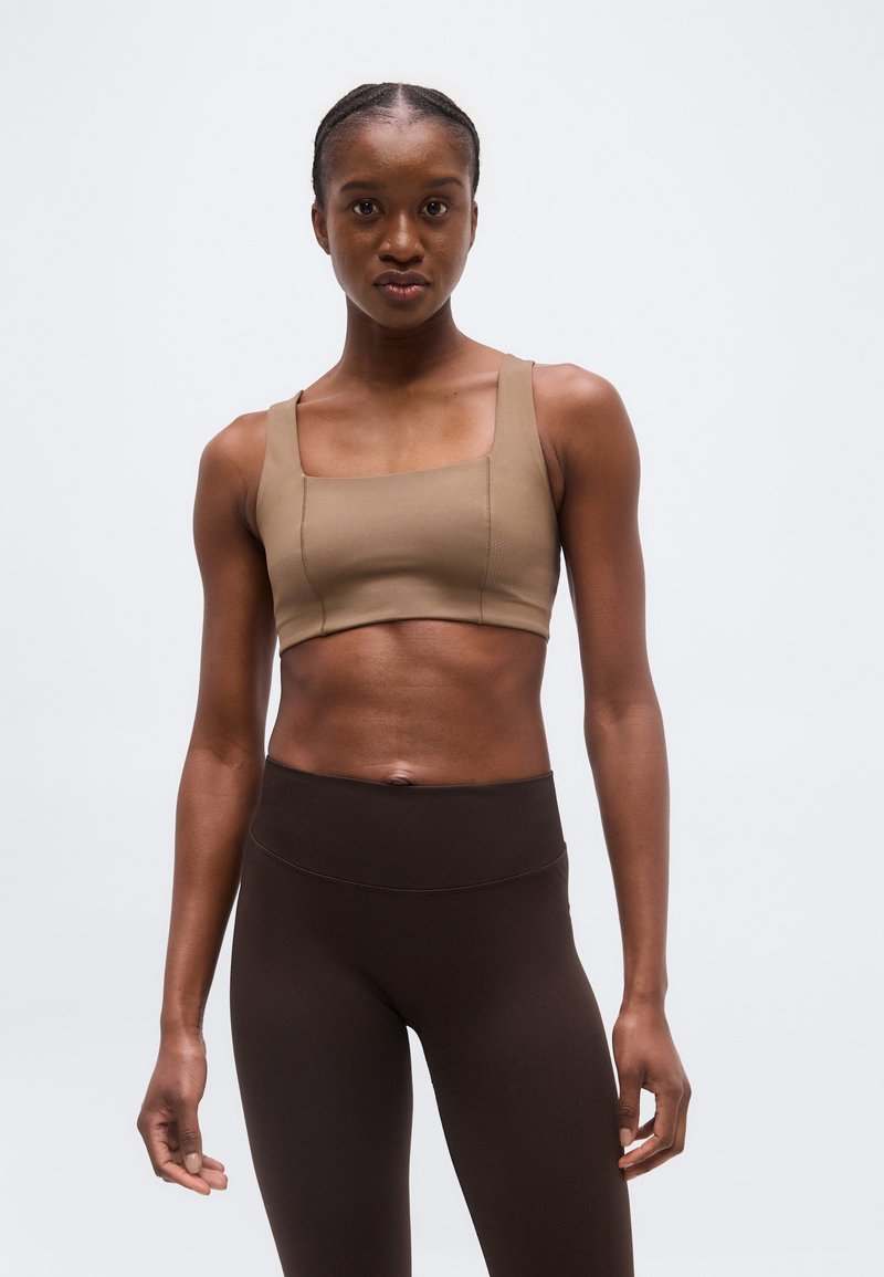 Γυναίκα όρθια, φοράει μπεζ κομμένη τοπ και ψηλόμεσα σκούρα καφέ κολάν, σε απλό φωτεινό φόντο.