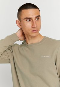 Lühikeste juustega mees kannab beeži svetti, millel on kiri "CASUAL FRIDAY", vaatab küljele, käsi puhkamas kaela taga.