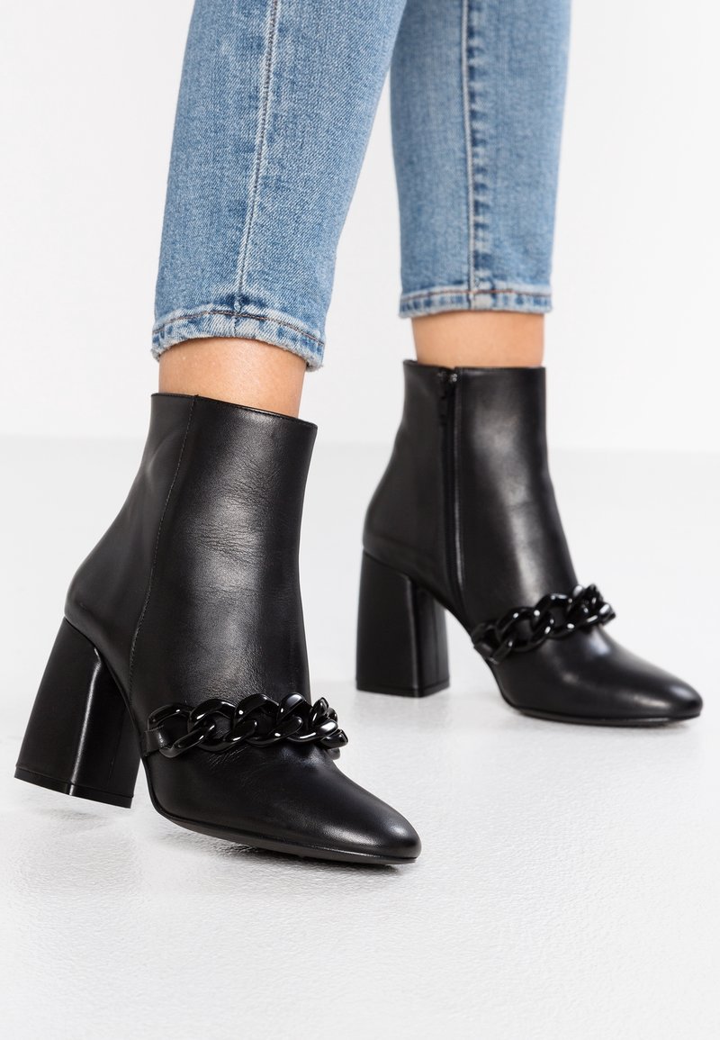 Botas de cuero negras hasta el tobillo con punta afilada, tacón de bloque y una cadena decorativa negra en la parte frontal. Acabado de superficie texturizada.