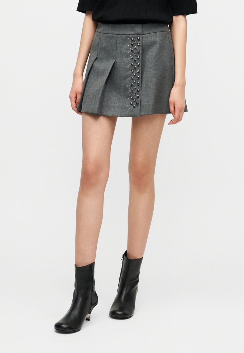 Γκρι πλισέ μίνι φούστα με μεταλλικά τρουκς κατά μήκος της αριστερής μπροστινής ραφής. Κατασκευασμένη από απαλό ύφασμα. Συνδυάζεται με μαύρα μποτάκια αστραγάλου.