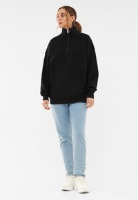 Felpa nera con colletto alto e mezza zip, dotata di tasche frontali e maniche ampie. Abbinata a jeans azzurri e sneakers bianche.