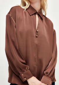 Brun silke bluse med krave, nøglehulåbning og knapdetaljer. Har lange, let puffede ærmer og elastiske manchetter.