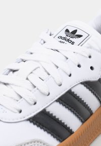 Biały skórzany sneaker z szarymi paskami, z teksturowaną gumową podeszwą, białymi sznurowadłami i czarną metką z logo na języku.