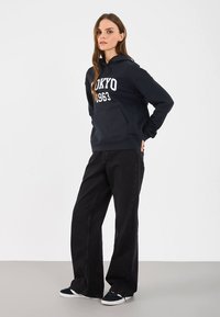 Γυναίκα που στέκεται φορώντας ένα μαύρο hoodie "Tokyo 1963", φαρδύ παντελόνι μαύρου χρώματος και μαύρα αθλητικά παπούτσια, μπροστά σε λευκό φόντο.