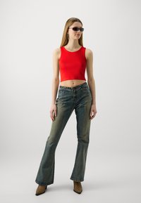 Rød ribbet croppet topp, høyhalsede falmede denim flared bukser, brune spisse ankelstøvletter og mørke solbriller. Minimalistisk bakgrunn.
