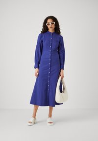 Abito blu con bottoni e maniche lunghe, caratterizzato da dettagli ricamati, silhouette dritta e lunghezza midi. Abbinato a accessori bianchi.