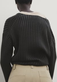 Personne portant un pantalon beige et un pull côtelé noir tombant sur une épaule, par-dessus un haut côtelé clair, vue de dos.