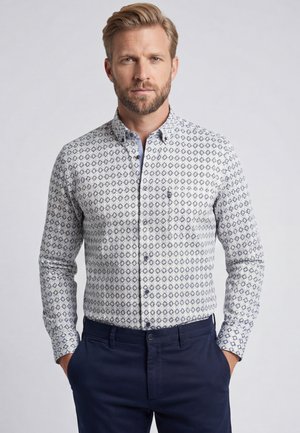 Man met baard die een witte blouse met lange mouwen met blauwe geometrische patronen en donkerblauwe broek draagt, staand met handen in de zakken.