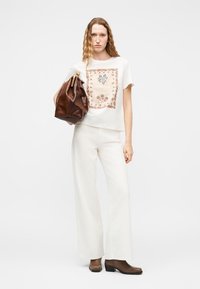 Γυναίκα με κυματιστά μαλλιά φοράει λευκό μπλουζάκι με λουλουδάτο μοτίβο, λευκό παντελόνι με φαρδύ πόδι, καφέ μποτάκια μέχρι τον αστράγαλο, κρατώντας μεγάλη καφέ δερμάτινη τσάντα.