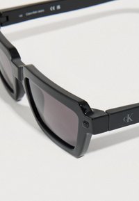 Μαύρα ορθογώνια γυαλιά ηλίου με σκούρους φακούς, διαθέτοντας γυαλιστερό πλαίσιο και μεταλλικές λεπτομέρειες. Το λογότυπο της μάρκας εμφανίζεται διακριτικά στο πλευρό.