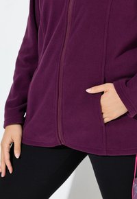 Persona che indossa una giacca in pile viola con cerniera, con una mano in tasca e leggings neri con una striscia rosa di lato.