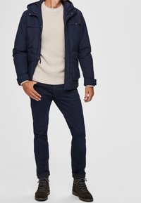 Tmavě modrá bunda s kapucí, béžový svetr, tmavé džíny a hnědé turistické boty. Bunda má dvě přední kapsy a zipové zapínání.