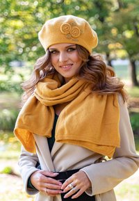 Geel baret met bloemenaccenten en bijpassende oversized sjaal, beide gemaakt van zachte stof; gedragen over een crèmekleurige jas tegen een vervaagde groene achtergrond.
