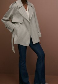 Zwart-witte geblokte trenchcoat met brede revers en riem, gedragen over een lichte top en donkere flared jeans. Soepele textuur, getailleerde pasvorm.