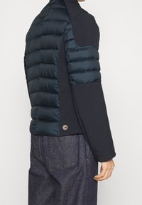 Marinjacka med en puffig, vadderad baksida, släta ärmar i tyg och en cirkulär logotypdetalj vid nederkanten.