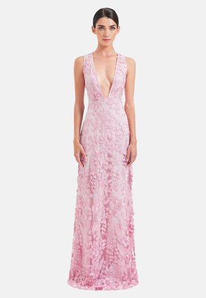 Donna che indossa un abito rosa senza maniche lungo fino a terra, con scollo profondo a V e applicazioni in pizzo floreale, in piedi davanti a uno sfondo bianco semplice.