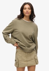 Felpa verde oliva con scollo a coste e logo discreto, abbinata a una gonna cargo beige chiaro con tasche laterali e dettagli con bottoni.