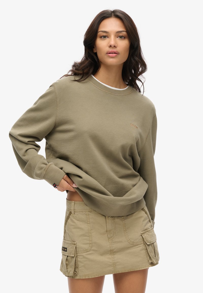 Felpa verde oliva con scollo a coste e logo discreto, abbinata a una gonna cargo beige chiaro con tasche laterali e dettagli con bottoni.