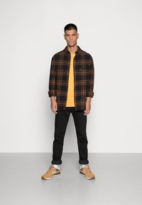 Camicia di flanella a quadri neri e marroni sopra una t-shirt color senape, abbinata a jeans neri e sneakers color beige, in piedi su uno sfondo grigio.