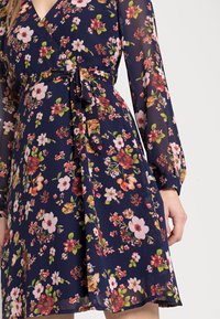 Vestido em tecido azul marinho, com mangas longas, cintura ajustada com um cinto amarrado e uma mistura de padrões florais em tons de rosa, vermelho e verde.