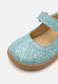 Zapato Mary Jane de purpurina turquesa con punta redondeada, tira de velcro y suela de goma. Presenta un acabado texturizado y brillante.