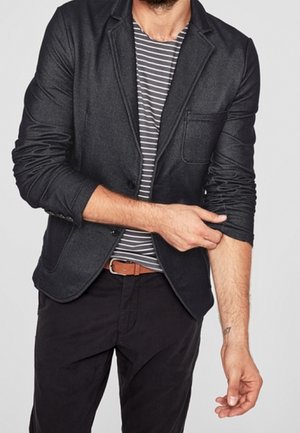 Mężczyzna podwijający rękawy ciemnoszarej marynarki, ubrany w pasiastą szarą i białą koszulę, czarne spodnie oraz brązowy pasek na jednolitym tle.