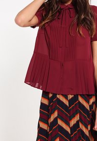 Blouse plissée bourgogne à manches courtes, col à nouer et plis verticaux ; associée à une jupe à motif chevrons multicolore.
