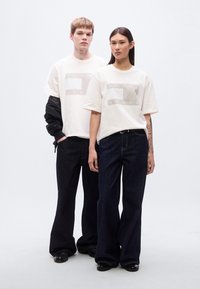 Λευκά γραφιστικά t-shirts με την επιγραφή "TOMMY JEANS", συνδυασμένα με φαρδιά σκουρόχρωμα τζιν παντελόνια. Ένα άτομο φοράει μαύρο μπουφάν πάνω από το πουκάμισό του.