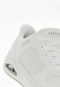 Białe Skechers Slip-Ins z gładką syntetyczną cholewką, perforowanymi akcentami, płaskimi białymi sznurówkami oraz wkładem amortyzującym z widoczną poduszką powietrzną.