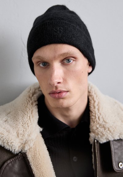 Berretto a maglia nero con finitura testurizzata, dotato di un logo sottile. Abbinato a una giacca con colletto in shearling marrone e camicia scura.