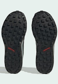 Suelas de goma negra con tacos multidireccionales, que presentan un patrón texturizado y el logo rojo "TRAXION", diseñadas para ofrecer tracción y agarre.