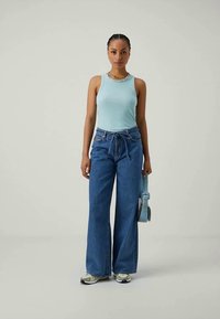 Top sin mangas de color azul claro, vaqueros de tiro alto azules con un detalle de lazo y zapatillas con acentos verdes, combinados con un bolso de hombro azul claro.