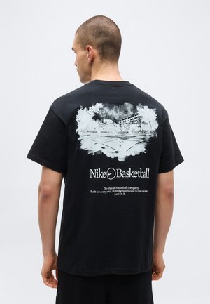 Hombre de espaldas vistiendo una camiseta negra de Nike Basketball con un gráfico blanco de una cancha de baloncesto al aire libre y árboles.