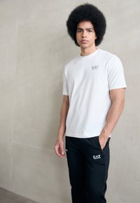 Fehér, rövid ujjú póló, amelyen egy kis EA7 logó található, fekete sportnadrággal párosítva, mindkettő sima anyagból készült. Minimál stílusú design.