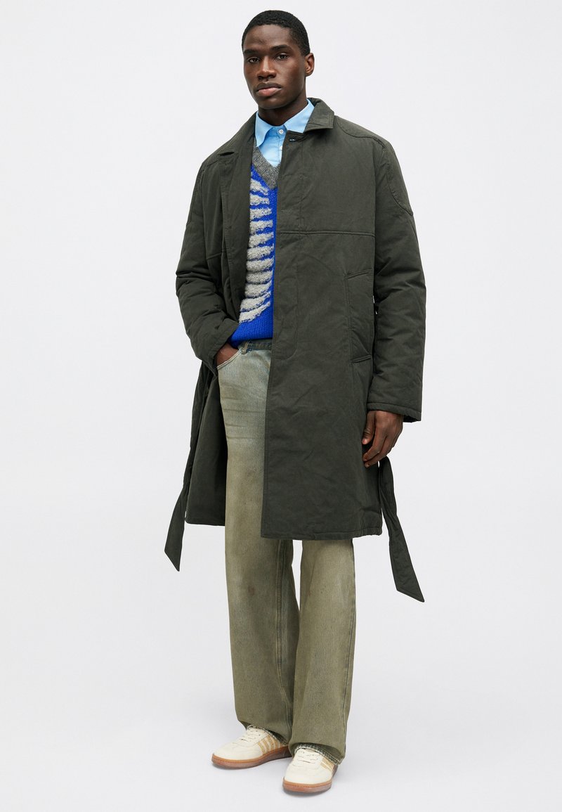 Olijfgroen mid-lengte jas met een kraag halslijn. Gepaard met een blauw patroontrui, een lichtblauwe shirt en lichtgroene broek.