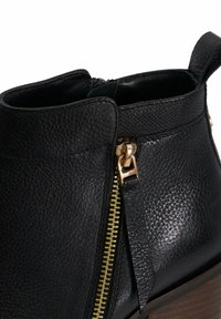 Zwarte leren enkellaars met een gestructureerde afwerking, voorzien van een gouden ritsdetail en een bruin houten hak. Accent is een zijhandvat.