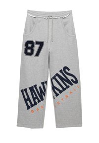 Szürke melegítőnadrág, húzózsinórral ellátott derékrésszel, oldalsó zsebekkel, navy kék grafikai "87" és "HAWKINS BASKETBALL" felirattal, narancssárga árnyalatokkal.