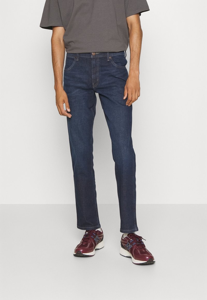 Homme portant un jean droit bleu foncé, un t-shirt gris, des chaussettes blanches et des chaussures de sport bordeaux, debout devant un fond uni.