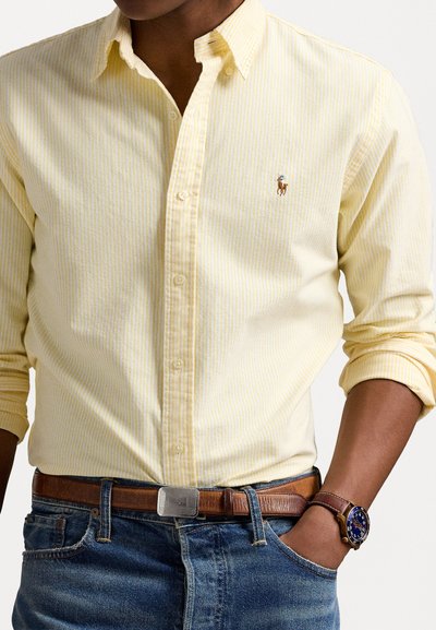 Camisa de botão amarelo claro com finas riscas brancas, apresentando um pequeno logótipo bordado. Usada com um cinto de couro castanho e um relógio azul.