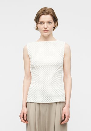 Femme portant un haut sans manches blanc texturé et un pantalon beige, debout devant un fond gris clair uni.