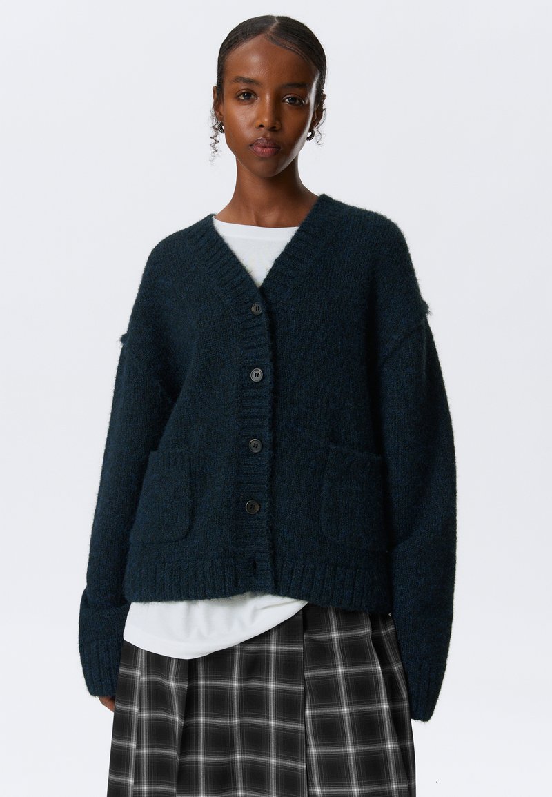 Dunkelgrüner Strickcardigan mit tiefem V-Ausschnitt, sechs schwarzen Knöpfen, zwei vorderen Taschen und gerippten Bündchen, über einem weißen Hemd und einem karierten Rock getragen.