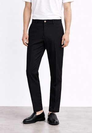 Mężczyzna ubrany w czarne, wąskie spodnie garniturowe oraz czarne skórzane mokasyny, stojący na tle jednolitej białej ściany.