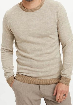 Mann trägt beigen strukturierten Langarmpullover mit gerippten Bündchen und Saum, kombiniert mit hellbrauner Hose, Hand in der Tasche ruhend.